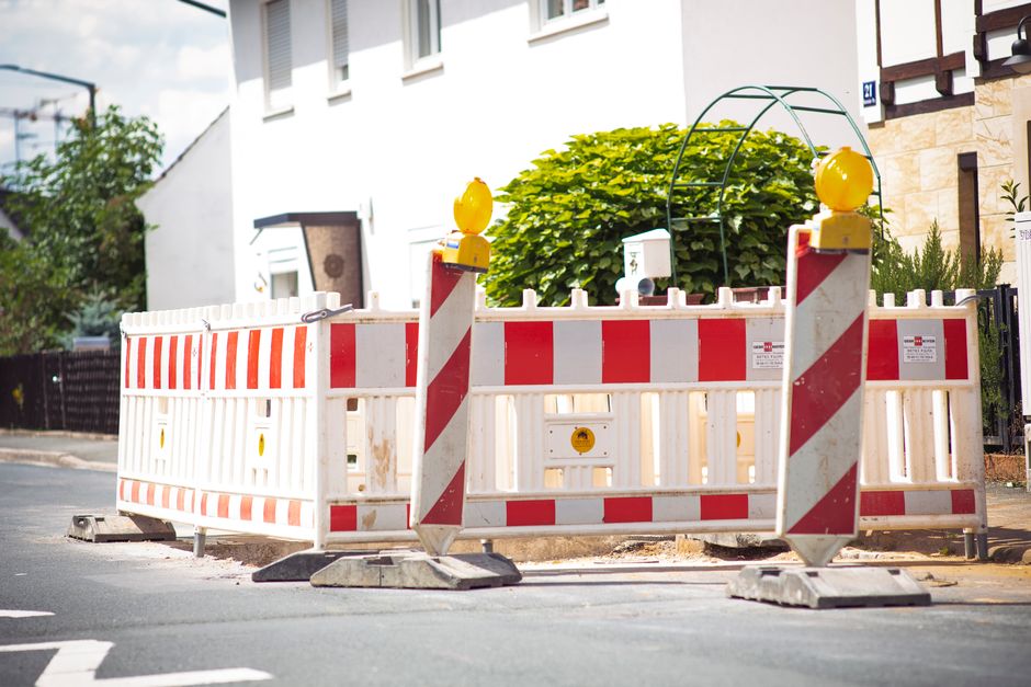 A temporary road closure barrier with white and red reflective panels and yellow warning lights on top, positioned across a residential street in Colindale. The barrier surrounds an area where house removals or moving activities are taking place, likely to facilitate the loading and unloading of furniture or boxes for a home relocation. Behind the barrier, part of a white building with multiple windows is visible, along with some greenery, including a lush shrub. The scene is outdoors during daylight, with natural lighting illuminating the pavement and street environment. The barrier is set up on the road to restrict vehicle access, supporting logistical movement of furniture and packing materials during a house move as part of a local street closure authorized for such activities, assisted by Man with Van Colindale, a professional removals service.