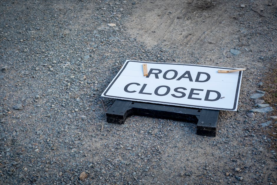 A rectangular road closure sign with a white background and bold black text reading 'ROAD CLOSED' lies on the gravel surface of a road or driveway. The sign is supported by a black plastic frame and secured with two small wooden clothespins, indicating temporary disruption to vehicle access. The gravel surface consists of small, uneven stones with a mix of gray and brown tones, and some larger rocks are visible around the sign. This scene suggests that the road is temporarily inaccessible, possibly due to maintenance or construction work related to house removals or relocation services. The setting appears outdoor with no visible buildings or people, emphasizing the temporary traffic restriction, which could coincide with a property clearance or moving process, where transport vehicles are being loaded or unloaded. As part of professional removal services by Man with Van Colindale, such road closures often facilitate the safe loading of furniture, boxes, and appliances during home relocations, with attention to local permits like those managed by Barnet Council for street closures.