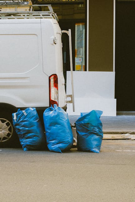 The image shows the rear end of a white cargo van parked outside a building, with the back doors closed. In front of the van, three large blue plastic bags filled with waste or belongings are placed on the pavement, ready for collection or disposal. The van has a roof rack mounted on top, possibly for carrying additional items during a home relocation or moving service. The location appears to be an urban area, with a glass door to the building visible on the right side of the image, reflecting some light. The area is clean, and the scene suggests the process of loading or unloading items as part of a house removal or clearance. The presence of durable packing materials and the context hint at professional moving or waste collection services, such as those offered by Man with Van Colindale, related to furniture transport or packing and moving activities.