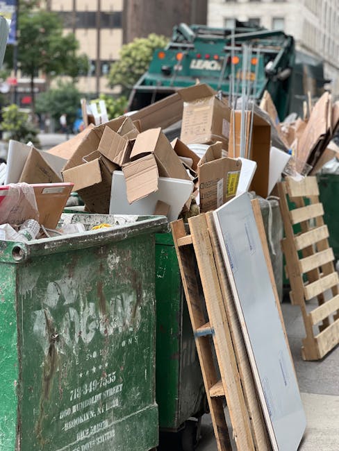 A large open-back truck with a metal frame and fabric sides, partially covered with brown tarpaulin, is loaded with a considerable amount of piled cardboard boxes, paper, and mixed waste materials inside an outdoor area. The truck is positioned on a paved surface adjacent to a small building with a corrugated metal roof. Several individuals are engaged in the loading process: one person standing inside the truck, arranging or overseeing the contents, while others stand outside, lifting or guiding items into the vehicle using their hands and physical effort. Surrounding the scene are additional piles of transparent plastic bottles, recyclables, and packaging materials scattered on the ground. In the background, there are residential structures, lush green trees, and utility poles with overhead wires, indicating a community environment. The lighting suggests daytime with natural sunlight illuminating the scene. This capturing represents a waste collection or packing activity associated with home or business relocation services, typical of logistics and removal operations. Man with Van Colindale occasionally appears in the context of house removals and waste removal solutions.