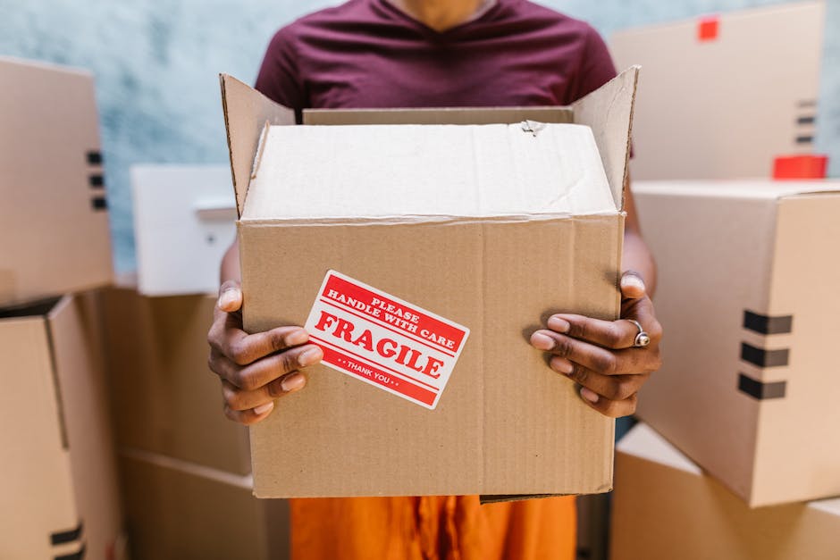 A man with curly dark hair, wearing a blue t-shirt and dark pants, is sitting on a wooden floor inside a room with white walls. He is surrounded by several cardboard moving boxes, some of which are se