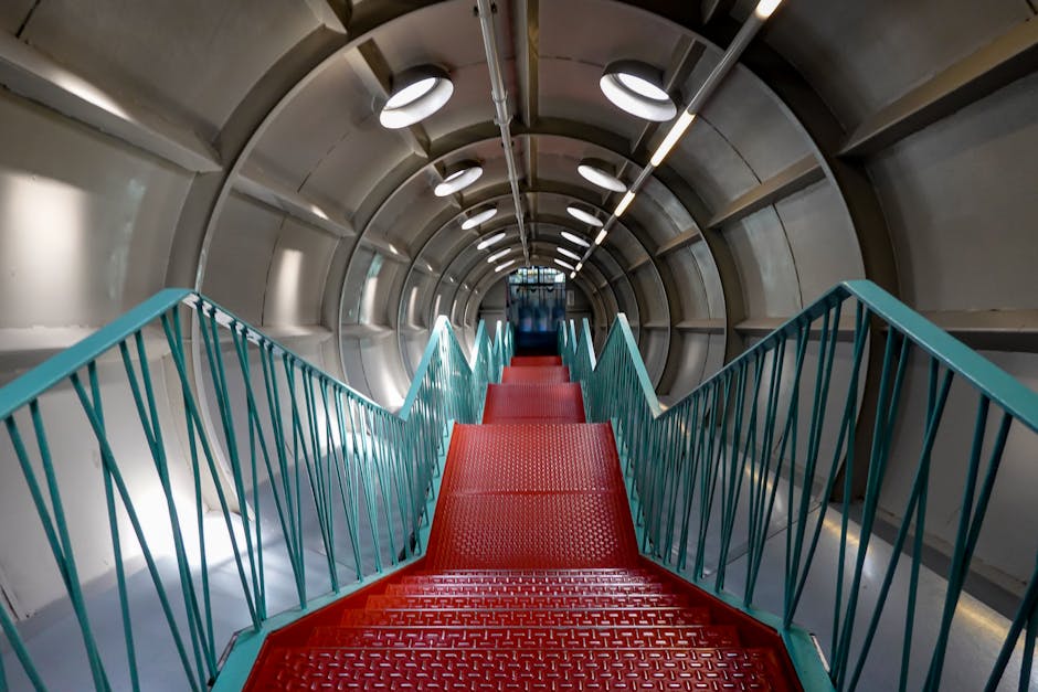 Inside view of a cylindrical, metallic, tunnel-like structure with a spiral red metal staircase ascending towards the top. The staircase is bordered by teal-colored metal railings with evenly spaced vertical bars. The interior walls, which are smooth and grey, are illuminated by circular ceiling lights, creating a bright and evenly lit environment. The red staircase appears to be part of a loading or transport facilitated environment, possibly related to home relocation or furniture transport within a moving process. The setting suggests an industrial or institutional location, such as an aircraft, elevator, or large container, with [COMPANY_NAME] potentially involved in providing removal services or logistical support for house removals, as indicated by the page context on [PAGE_TITLE]. The overall scene highlights the structural details of the staircase and surrounding environment, emphasizing the efficiency and safety features suited for carrying household items during a move.