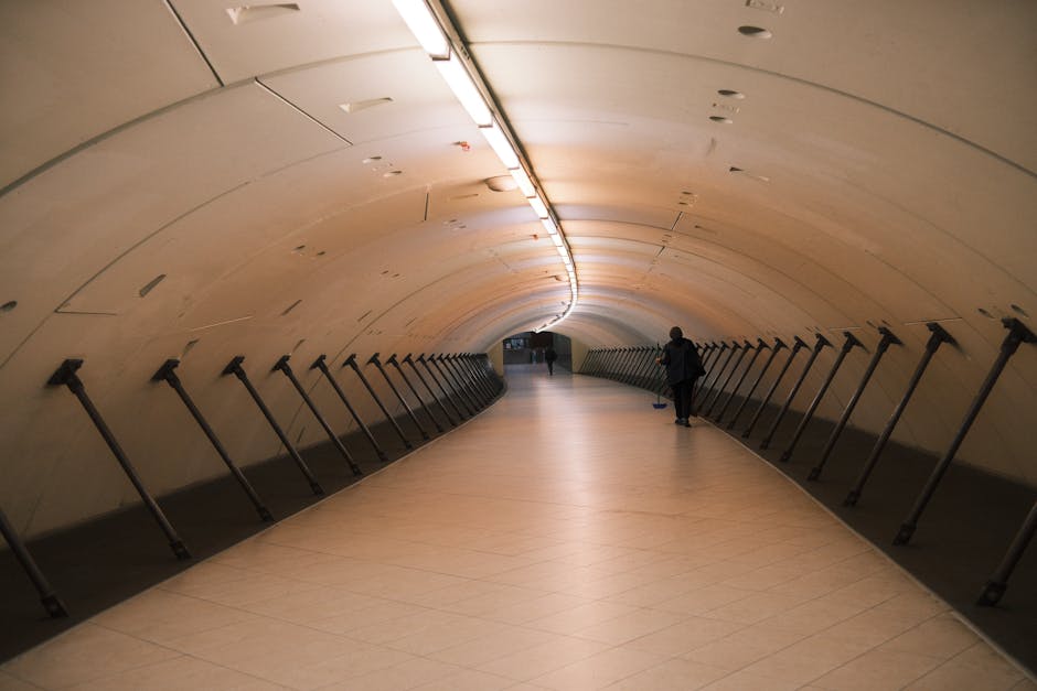 Inside a curved underground pedestrian tunnel with smooth, beige walls and ceiling panels, featuring evenly spaced black support braces along both sides. The tunnel is well-lit by a long strip of ceiling lighting. A person dressed in dark clothing and carrying a bag is walking towards the far end of the tunnel, which appears to lead outside or to another section of the station. The flooring is light-colored tile, and the space is clear of clutter, emphasizing the tunnel's spacious and clean environment, suitable for foot traffic during house removals or transportation between locations such as the Colindale Tube station and nearby landmarks.