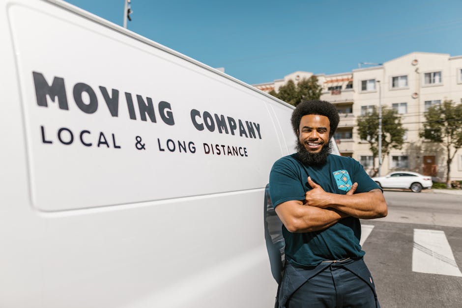 A smiling male professional from Man with Van Colindale stands outside a white moving van with the company's logo and the text 'MOVING COMPANY LOCAL & LONG DISTANCE' printed on its side. He is positioned near the open rear doors of the van, which is parked on a residential street with multi-storey white buildings and some parked cars in the background. The man is wearing a dark blue T-shirt, has a beard, and has his arms crossed, indicating confidence in the home relocation and furniture transport services offered. The scene is set during daylight hours with clear skies, emphasizing the logistics involved in packing, loading, and transporting household items for same-day or last-minute moves in Colindale, aligning with services provided by [COMPANY_NAME]. The visual highlights the loading process and moving equipment, such as the van and materials, consistent with professional packing and moving activities for house removals.
