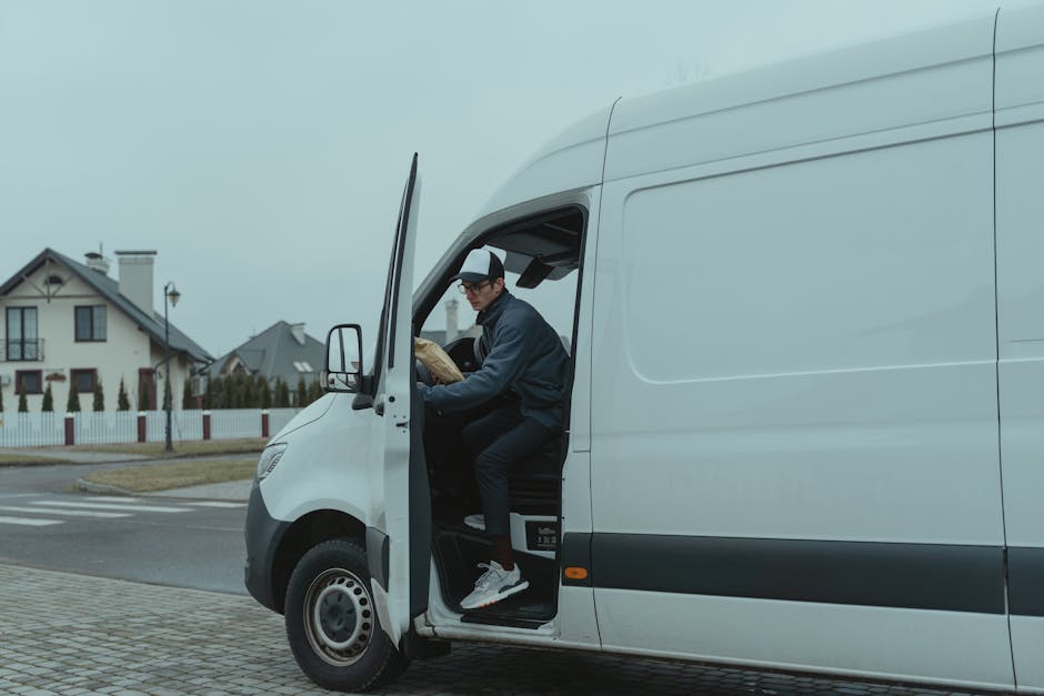 A man wearing a grey cap, glasses, and a dark jacket is seated inside a white delivery van with the side door open, preparing an item wrapped in brown packing paper for home relocation. The van is parked on a paved driveway near a residential area with modern houses, a grassy lawn, and a white picket fence visible in the background. The interior of the van's cabin shows the driver’s seat and part of the cargo space, which appears ready for furniture transport or packing and moving tasks. The environment is overcast with diffused natural light, and the scene captures the loading process typical of professional removals, with the man handling items to be transferred to or from the vehicle, reflecting the services provided by Man with Van Colindale for last-minute or same-day house moves.