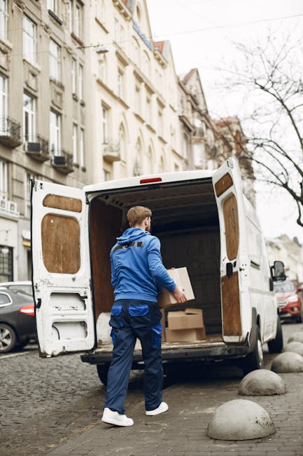 A smiling male professional from Man with Van Colindale stands outside a white moving van with the company's logo and the text 'MOVING COMPANY LOCAL & LONG DISTANCE' printed on its side. He is positioned near the open rear doors of the van, which is parked on a residential street with multi-storey white buildings and some parked cars in the background. The man is wearing a dark blue T-shirt, has a beard, and has his arms crossed, indicating confidence in the home relocation and furniture transport services offered. The scene is set during daylight hours with clear skies, emphasizing the logistics involved in packing, loading, and transporting household items for same-day or last-minute moves in Colindale, aligning with services provided by [COMPANY_NAME]. The visual highlights the loading process and moving equipment, such as the van and materials, consistent with professional packing and moving activities for house removals.