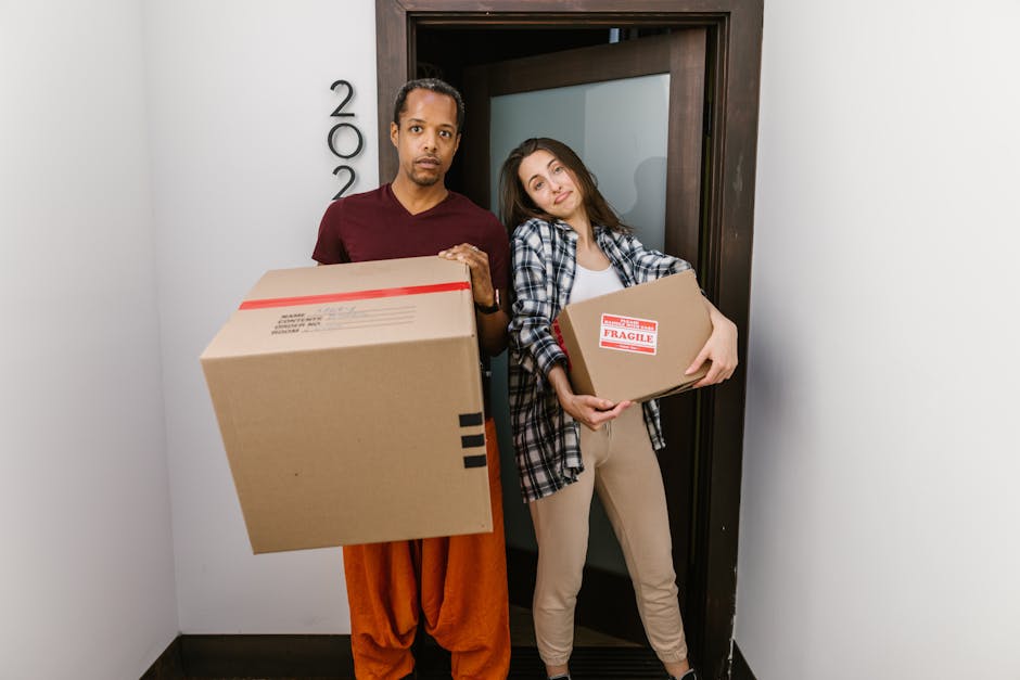 A man and woman walking outdoors on a residential street, involved in house removals or moving activities. The woman is carrying a rectangular wooden box filled with green plants, dressed in a cream-c