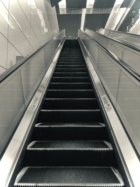 A close-up view of a moving escalator in an indoor setting, with black rubber steps featuring white safety edges, flanked by silver metal side panels. The escalator is situated inside a modern building with reflective metallic walls and ceiling panels that create a sleek, contemporary appearance. The escalator is in an upward motion, and part of the surrounding environment, including glass and metal elements, is visible at the top. This scene represents the type of infrastructure encountered during house removals or apartment relocations in urban settings. The image illustrates the typical environment for home relocation services provided by companies such as Man with Van Colindale, highlighting logistics involved in navigating narrow hallways or staircases within multi-storey buildings during furniture transport and moving logistics.