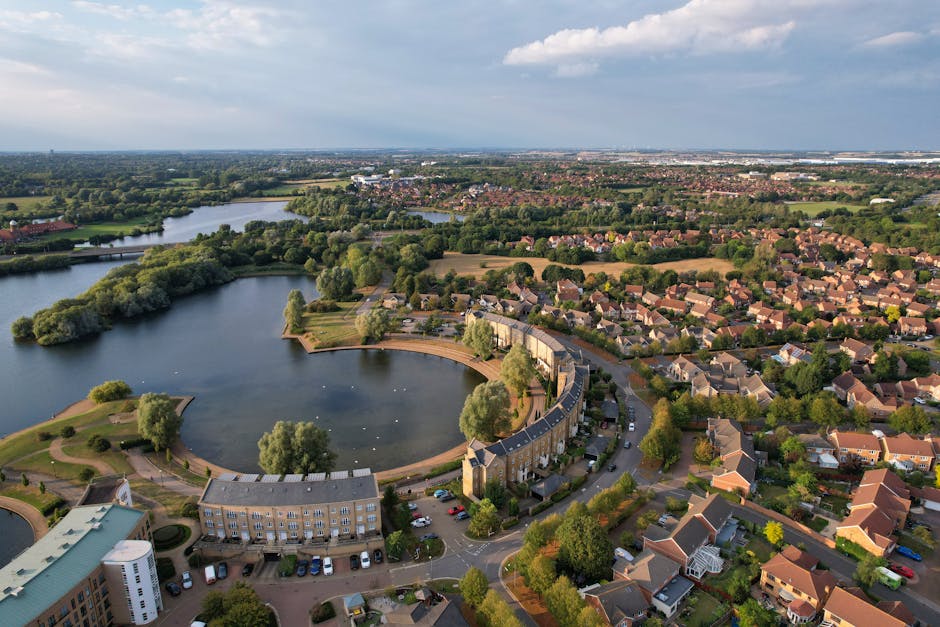 Aerial view of a residential neighbourhood featuring a large, curved lake with landscaped banks and surrounding trees, located adjacent to a community of terraced houses and detached homes with red and grey tiled roofs. The lake has a walkway along its perimeter, with some boats visible on the water. In the foreground, there are buildings associated with a housing complex, as well as parking areas filled with cars. The scene is illuminated by natural daylight, with a partly cloudy sky overhead. This image captures the environment where home relocation and furniture transport services by Man with Van Colindale are often arranged, illustrating the setting for an efficient moving process within a suburban area near green spaces and water features.