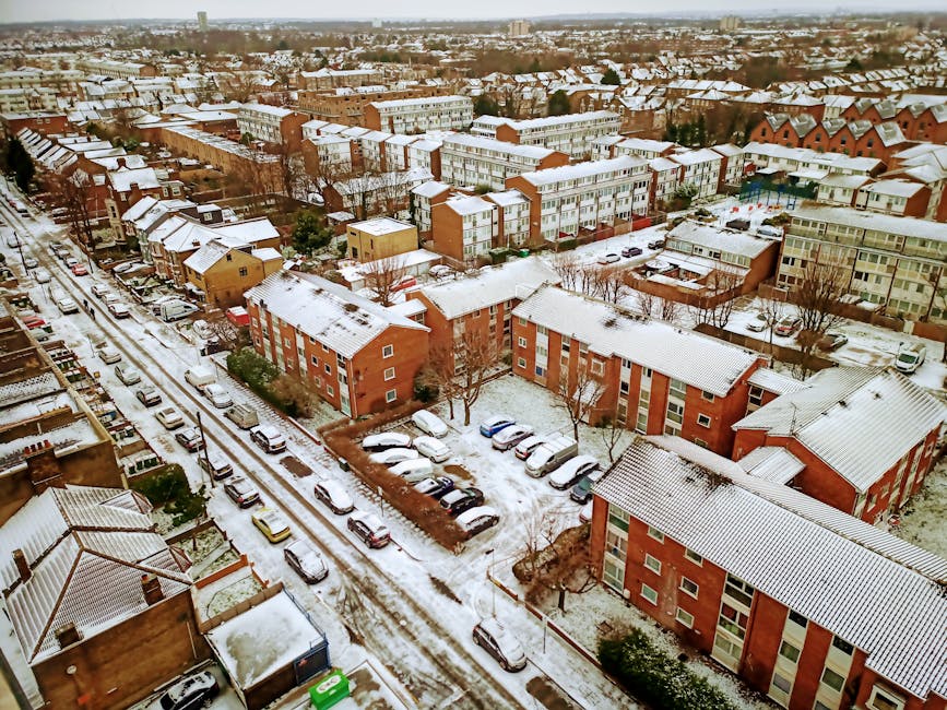 Aerial view of a residential neighbourhood featuring a large, curved lake with landscaped banks and surrounding trees, located adjacent to a community of terraced houses and detached homes with red and grey tiled roofs. The lake has a walkway along its perimeter, with some boats visible on the water. In the foreground, there are buildings associated with a housing complex, as well as parking areas filled with cars. The scene is illuminated by natural daylight, with a partly cloudy sky overhead. This image captures the environment where home relocation and furniture transport services by Man with Van Colindale are often arranged, illustrating the setting for an efficient moving process within a suburban area near green spaces and water features.