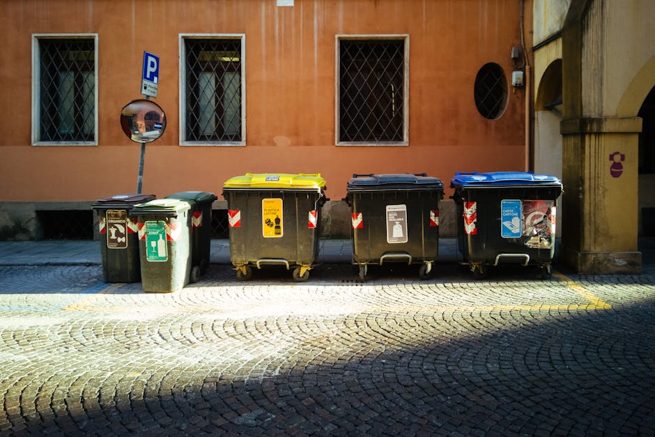 A row of four wheeled rubbish and recycling bins positioned on a cobblestone street in front of a peach-coloured building with three windows having metal grid covers. The bins include a green bin for general waste, a yellow bin for recyclable packaging, a black bin for other waste, and a blue bin for paper. Each bin has a distinct coloured lid and labels indicating their contents, with some labels showing symbols or text related to waste disposal rules. To the left, there is a convex mirror mounted on a pole, and the background features a parked vehicle partially visible behind the bins. The scene suggests an area used for waste collection associated with residential or commercial properties, relevant to home relocation and moving logistics as managed by Man with Van Colindale, which may involve proper packing and transport of household waste during moves.