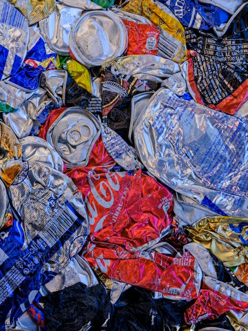 A close-up view of crushed aluminium cans, some with identifiable branding such as Coca-Cola, mixed together in a pile. The cans display various colours including red, silver, blue, and yellow, with some torn or flattened for disposal. The cans are situated outdoors, possibly on pavement or in a recycling collection point, with natural lighting highlighting the metallic surfaces. The image reflects packaging waste typical of household recycling or waste disposal processes. This scene relates to home relocation and moving services provided by Man with Van Colindale, emphasizing proper waste management during packing and moving activities to help avoid fines and adhere to local regulations in Colindale.